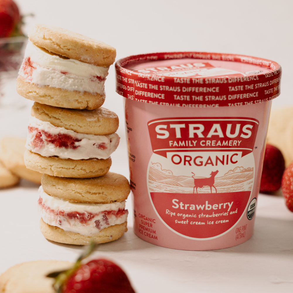Stack of strawberry ice cream sandwiches next to a Straus Family Creamery organic strawberry ice cream pint with fresh strawberries.